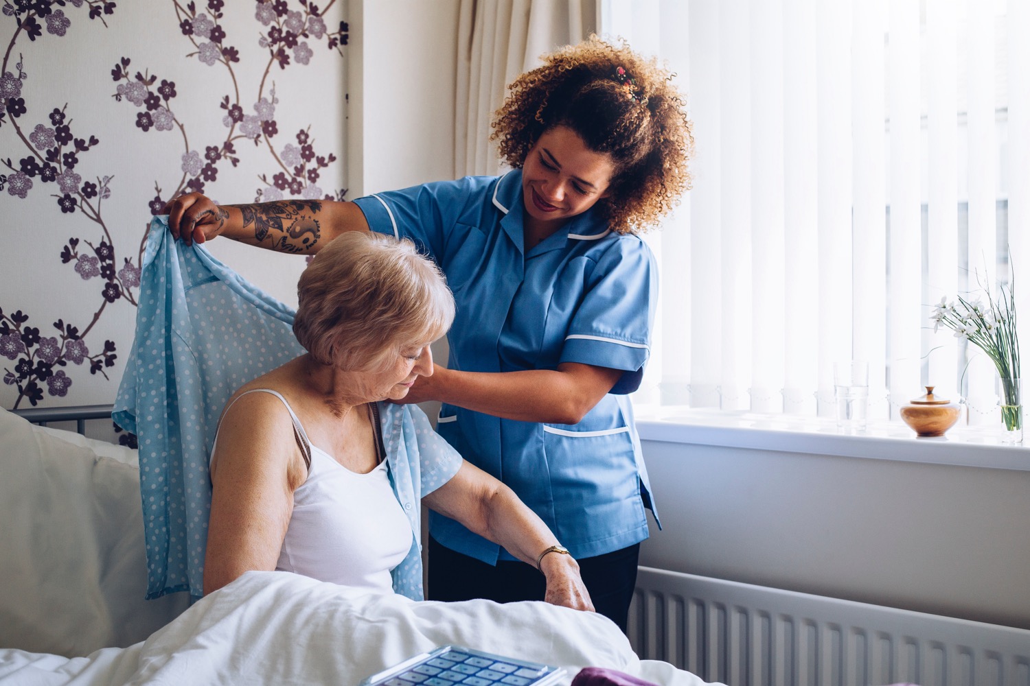 An African American caregiver with arm tattoos helps dress an elderly woman receiving in-home care.